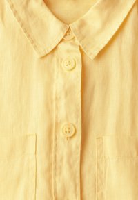 Camicia di lino gialla con colletto, cinque bottoni gialli e due tasche sul petto. Texture morbida con un aspetto leggero e traspirante.