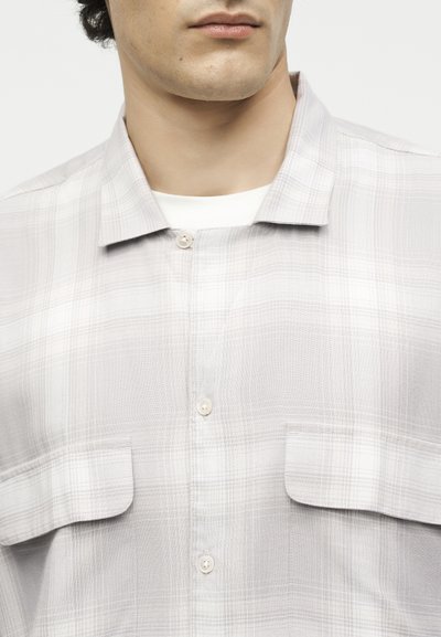 Άνδρας που φοράει ένα ανοιχτογκρίζο καρό πουκάμισο με κουμπιά πάνω από ένα λευκό μπλουζάκι με στρογγυλή λαιμόκοψη, εμφανίζοντας γιακά και τσέπες στο στήθος.