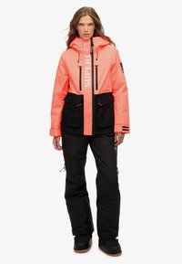 Jasnoróżowa wodoodporna kurtka z czarnymi akcentami, dwoma przednimi kieszeniami, regulowanym kapturem, w komplecie z czarnymi spodniami i butami.