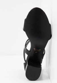 Sandale à talons pour femme noire montrant une semelle lisse avec l'inscription "minelli made in Brazil 37", appuyée contre un mur et une surface blancs.
