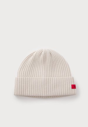 Berretto a maglia a coste color crema con risvolto e piccolo etichetta rossa sul lato, su sfondo bianco.
