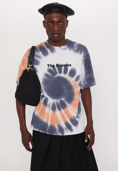 Balts, tumši zils un oranžs t-krekls ar tievdiezēšanas tehniku, uz kura redzams "The Kooples" logo melnā krāsā. Modelis valkā melnu somu un melnu bereti.