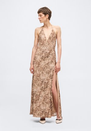 Kvinde iført en ærmeløs, halterneck maxi kjole med leopardprint og høj sidelågspræning, stående og kiggende sidelæns mod en ensfarvet baggrund.