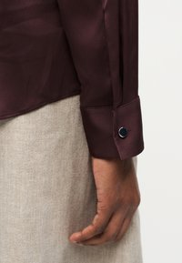 Καφέ μανσέτα πουκάμισου από σατέν με μαύρο κουμπί, φορεμένη πάνω από ανοιχτό μπεζ υφασμάτινο παντελόνι, με ένα χαλαρό χέρι να ξεκουράζεται προς τα κάτω.