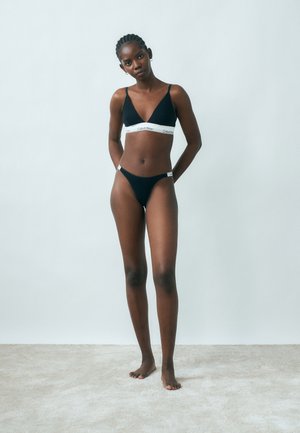 Vitka žena s pletenicama, odjevena u crni Calvin Klein grudnjak i donje rublje, stoji bosa na svijetlom tepihu, na jednobojnoj pozadini.