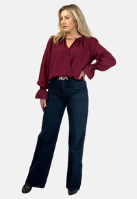 Bordowa bluzka z marszczonymi rękawami, zestawiona z ciemnoniebieskimi spodniami o szerokich nogawkach. Kobieta nosi srebrny naszyjnik i czarne szpilki.