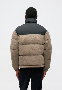 Geacă puffer cu secțiune superioară din nailon negru și secțiune inferioară dintr-un material texturat bej, cu coase vizibile și mansete elastice.