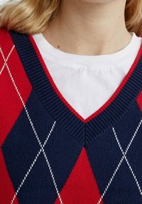 Blond persoon die een rood en marineblauw ruitjestrui zonder mouwen met witte diamantenpatronen over een eenvoudige witte ronde hals T-shirt draagt.