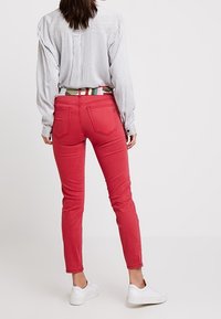Jean rouge ajusté avec une texture lisse, design à cinq poches et une ceinture à motif contrasté. Associé à une chemise rayée et des baskets blanches.