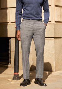 Pantaloni grigi intrecciati con una vestibilità sartoriale, dotati di tasche laterali e una texture liscia. Abbinati a scarpe in pelle nere e a un maglione blu.