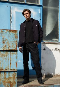 Giacca in tela blu marina sopra una t-shirt nera, abbinata a pantaloni neri e scarpe nere. Il modello è posato su uno sfondo industriale blu.