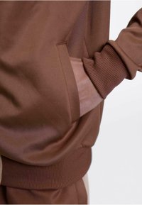Brun idrettsgenser med ribbet mansjett og lommebeskrivelse. Glatt tekstur og tettsittende design som viser en moderne silhuett.