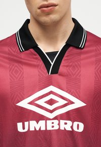 Približnji posnetek osebe, ki nosi maročno športno majico Umbro s črnim ovratnikom in belim logotipom na prsih.
