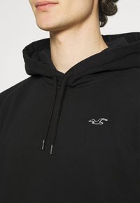 Hombre vestido con una sudadera negra con cordones y un pequeño logo de un pájaro blanco en el lado izquierdo del pecho, sobre un fondo liso.