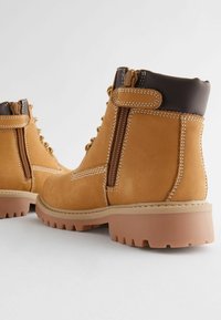 Tan mockasins med en brun läderkrage, med sidodragkedjor, snörning och en tjock gummisula för bra grepp.