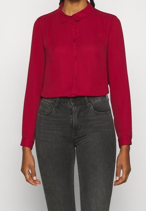 Mujer que lleva una blusa roja de manga larga abotonada metida en unos vaqueros de tiro alto de color gris oscuro sobre un fondo liso.