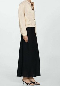 Kobieta ubrana w beżową kurtkę z guzikami, długą czarną spódnicę oraz czarne sandały na szpilkach, stojąca na tle jasnego, jednolitego tła.