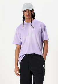 Camiseta de algodão roxa com corte descontraído, mangas curtas e gráfico discreto. Combinada com calções pretos com bolsos cargo e cordão de ajustamento.