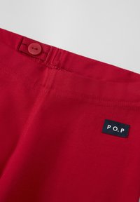 Punaiset puuvillaiset housut, joissa on sileä pinta, vyötärö, jossa on samansävyinen napitus, ja pieni musta etiketti, jossa on "P.O.P"-design.