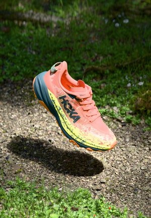 Hoka running shoe in bright coral and yellow, featuring a breathable mesh upper, thick cushioned sole, and prominent black logo.