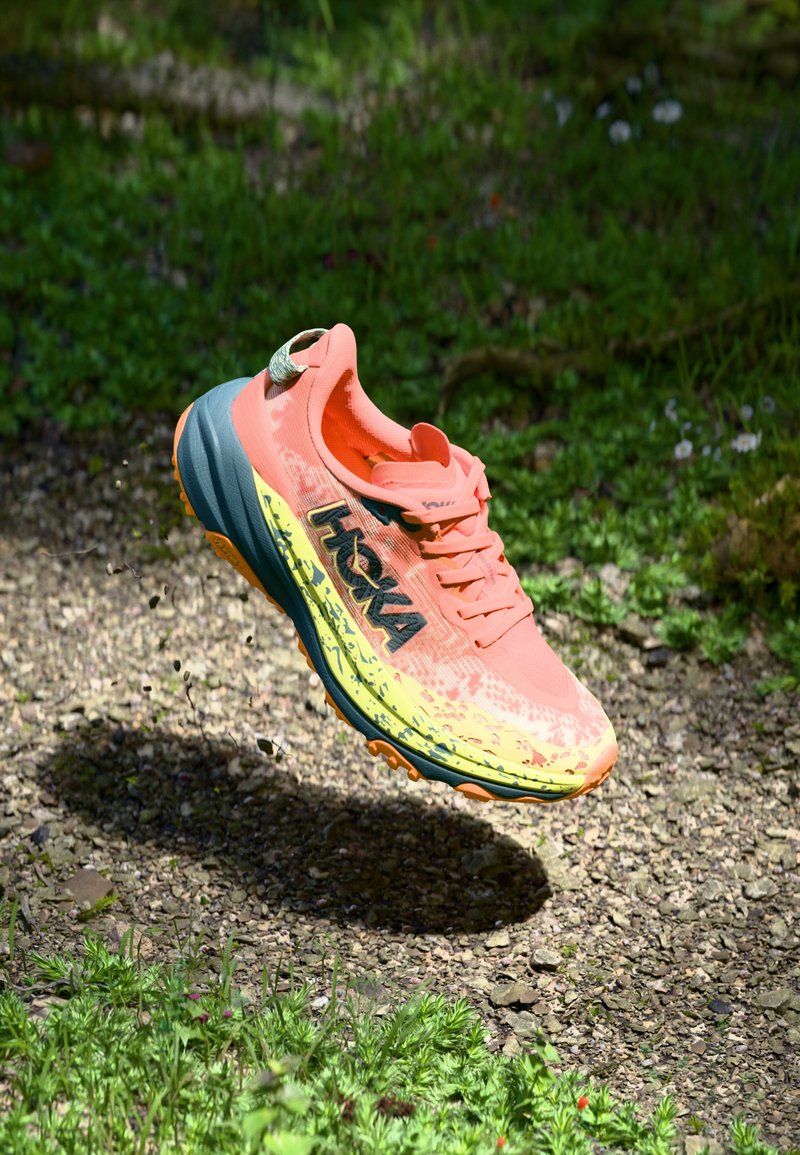 Hoka running shoe in bright coral and yellow, featuring a breathable mesh upper, thick cushioned sole, and prominent black logo.