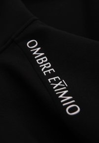 Tela negra con una textura suave, con el texto "OMBRE EXÍMIO" bordado en blanco, ubicado a lo largo de un lado.