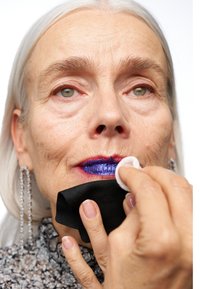 Gros plan sur une personne en train d'enlever un rouge à lèvres bleu vif avec un disque de coton. Possède des cheveux courts argentés, des rides visibles et de longues boucles d'oreilles en cristal.