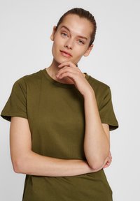 Olivengrøn bomulds T-shirt med korte ærmer, rund hals og glat tekstur. Modellen poserer med den ene hånd på hagen.