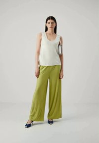 White sleeveless top, pleated olive green wide-leg pants, and blue heeled shoes. Model holds a silver clutch, standing against a neutral background.