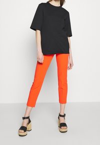 Černé oversized tričko, výrazné oranžové zkrácené kalhoty a černé platformové sandály s dřevěnými podpatky. Jednoduchý design; žádné viditelné vzory.
