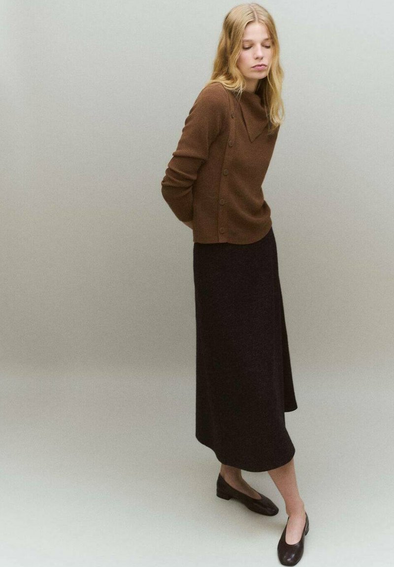 Pull en laine marron avec un col châle et des détails en boutons, associé à une longue jupe noire et des chaussures plates sombres sur un fond neutre.