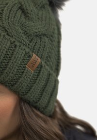 Gorra de punto verde con trenzas texturizadas y un pompón negro en la parte superior. Cuenta con un parche de logo de cuero marrón en el lado.