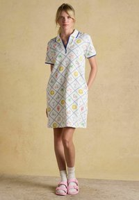 Blonde Frau trägt ein weißes Polo-Kleid mit kurzen Ärmeln und bunten Sommer-Ikonen, weiße Socken und rosa Sandalen, steht auf einem beigen Boden.