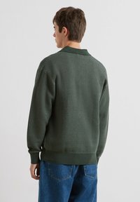 Zielony sweter z dzianiny o fakturowanym wzorze, z prążkowanymi mankietami i dołem, z kołnierzem. Noszony z niebieskimi dżinsami.