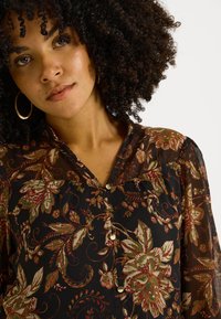Mujer con cabello rizado que lleva pendientes de aro dorados y una blusa negra abotonada con un patrón floral rojo y verde sobre fondo blanco.