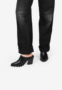 Mules de cuero negro con punta afilada y tacón cuadrado. Decorados con tachuelas plateadas en la parte delantera. Llevados con vaqueros negros enrollados.