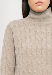 Frau in einem beigen, kabelstrickartigen Rollkragenpullover, der eine detaillierte Textur und ein Muster zeigt, vom Mund bis zu den Schultern zugeschnitten.