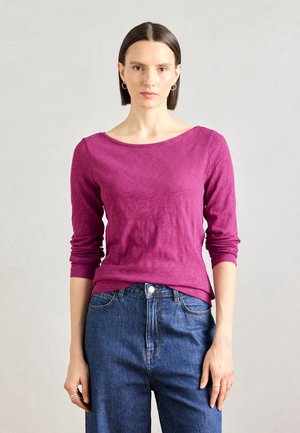 Langermet topp med scoop-neck i teksturert magenta stoff, kombinert med høyt liv blå denimjeans. Diskré sømdetaljer på ermene.