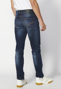 Sötétkék farmernadrág, slim fit szabással, két hátsó zsebbel és enyhe kopással. Fekete derékpánt texturált részlettel. Fehér sportcipővel viselve.