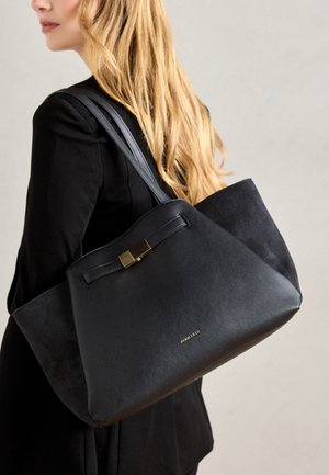 Lyshåret kvinde i sort outfit bærer en stor sort taske med guldlås og "ANNA FIELD"-logo på skulderen.