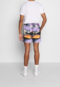 Kolorowe szorty tie-dye w fioletowe, pomarańczowe i czarne wzory. Elastyczny pasek w talii i dwie boczne kieszenie. Noszone z białymi sneakersami i skarpetkami.