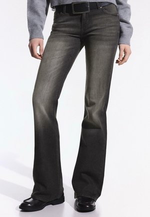 Frau trägt ausgewaschene schwarze Bootcut-Jeans, schwarzen Gürtel, grauen Pullover und schwarze Schuhe, steht mit entspannt an den Seiten hängenden Händen.