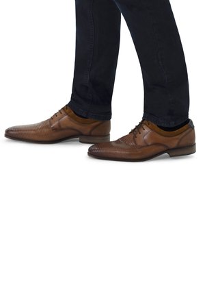 Chaussures en cuir marron avec un motif texturé, dotées d'un bout capé et d'accents décoratifs, associées à un jean foncé. Semelle lisse et lacets.