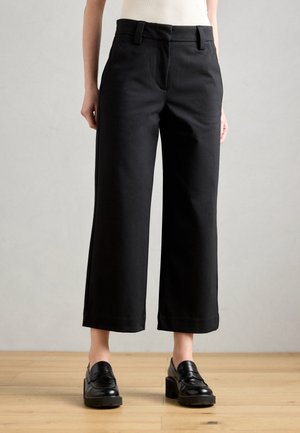 Person wearing black wide-leg cropped trousers and black heeled loafers standing on wooden floor against light gray wall.