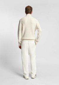 Maglione cream a trecce con colletto alto, abbinato a pantaloni bianchi. Il tessuto presenta un motivo a zigzag testurizzato e un orlo a coste.