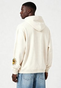 Felpa con cappuccio color crema a vestibilità rilassata. Presenta un design di sole ricamato sulla manica sinistra. Texture morbida e polsini a costine.