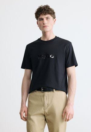 Νεαρός άνδρας με σγουρά μαλλιά, φορώντας μαύρο κοντομάνικο μπλουζάκι με το κείμενο "HUGO" και μπεζ παντελόνι, μπροστά σε απλό ανοιχτό φόντο.