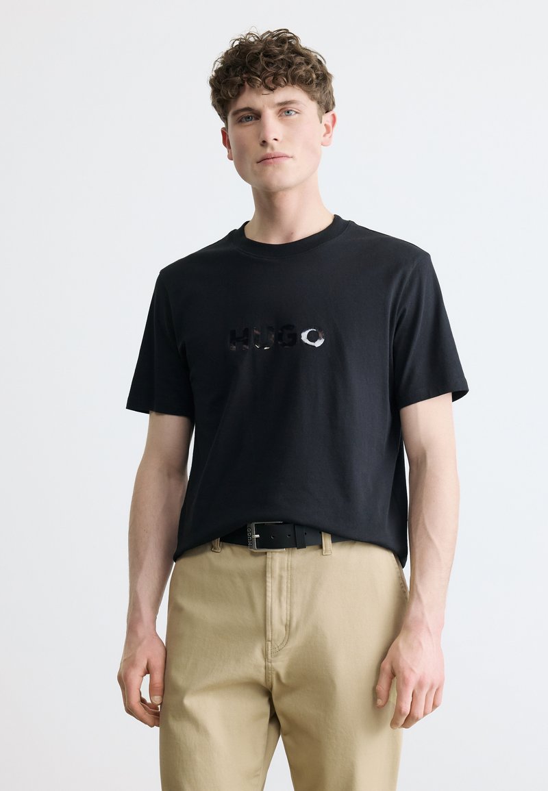 Νεαρός άνδρας με σγουρά μαλλιά, φορώντας μαύρο κοντομάνικο μπλουζάκι με το κείμενο "HUGO" και μπεζ παντελόνι, μπροστά σε απλό ανοιχτό φόντο.