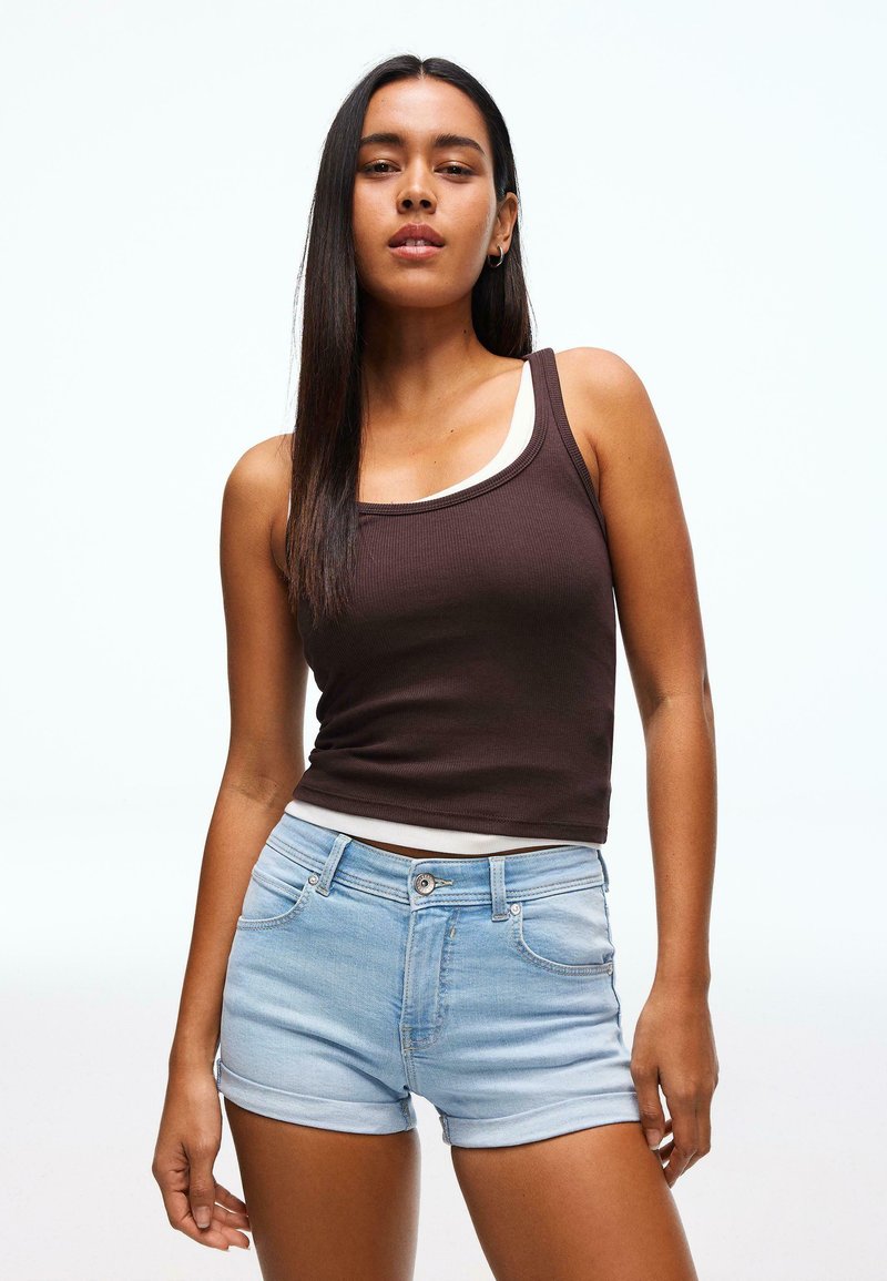 Hnedé rebrované tielko vrstvené cez biele tielko, doplnené svetlomodrými rifľovými šortkami so zvinutými lemami a prednými vreckami.