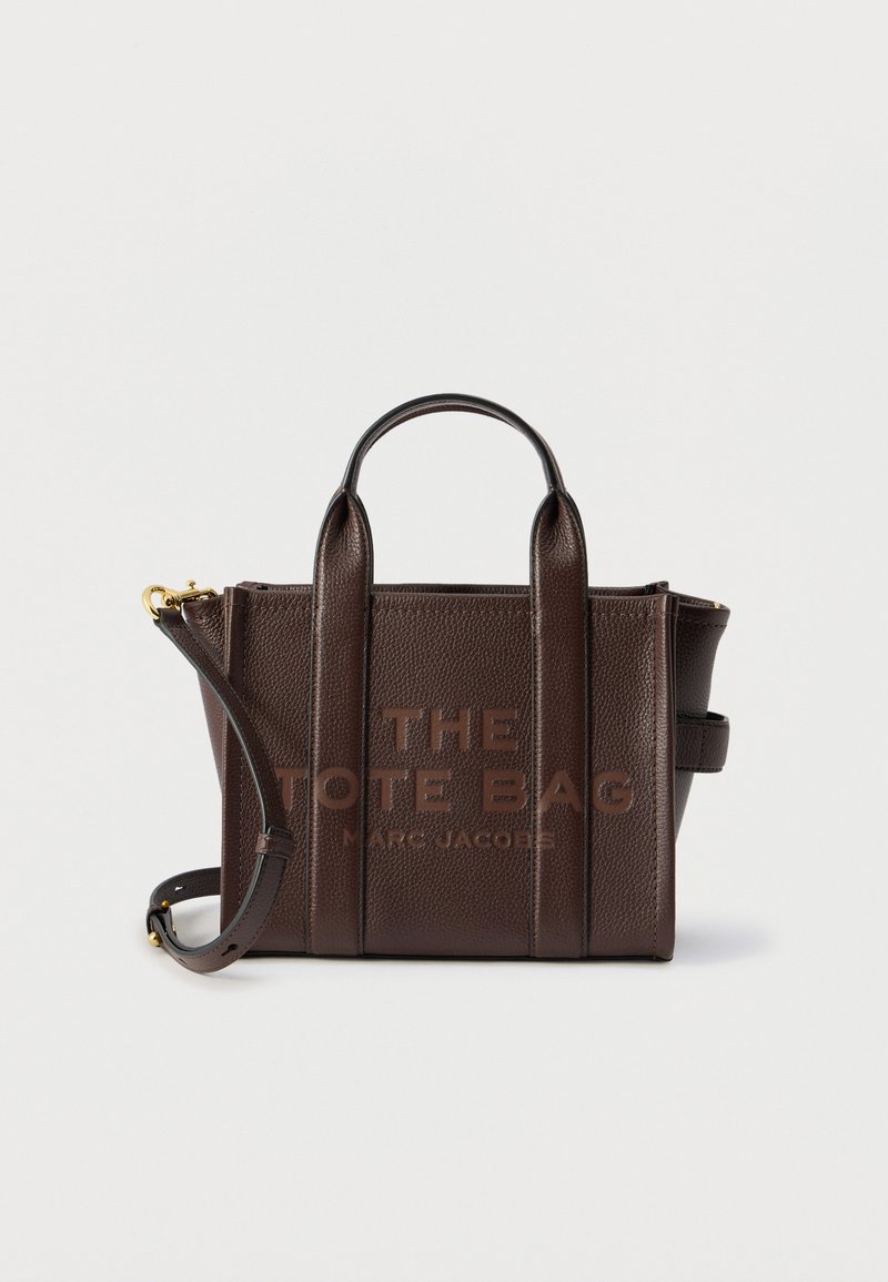Τσάντα tote από σκουρό καφέ δέρμα με υφή πετρωμάτων, με επάνω λαβές, αφαιρούμενο ιμάντα και ανάγλυφο κείμενο "THE TOTE BAG MARC JACOBS" στο μπροστινό μέρος.